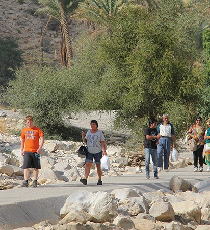 Oman Adventure Tour group walking along a rocky valley path surrounded by palm trees and desert hills in Oman.