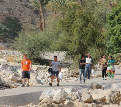 Oman Adventure Tour group walking along a rocky valley path surrounded by palm trees and desert hills in Oman.