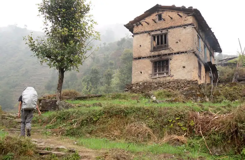 Trekker approaching a traditional stone house in Simigaun village, surrounded by misty hills and lush greenery.