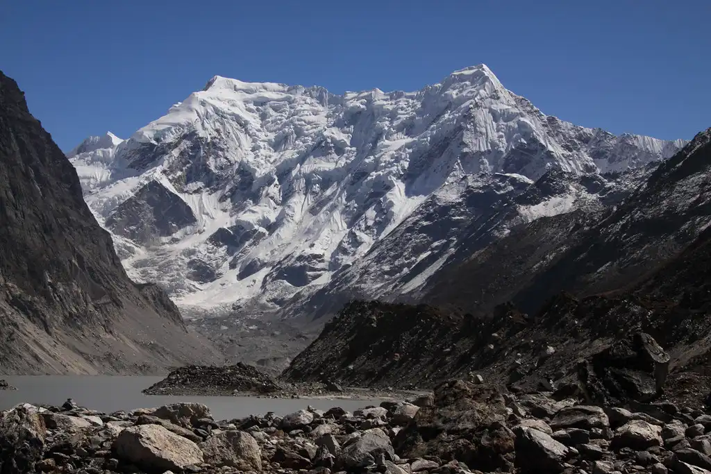 Tso Rolpa Lake with snow-covered Himalayan peaks in the background, surrounded by rugged rocky terrain.
