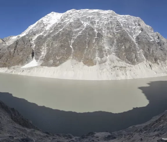 Panoramic view of Tso Rolpa glacial lake surrounded by snow-capped peaks with trekkers on the trail.