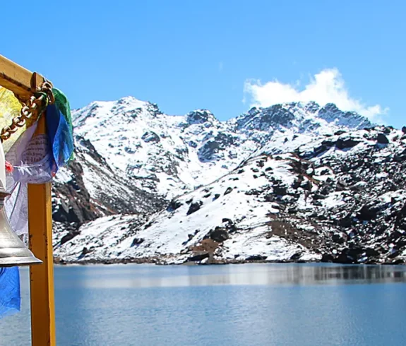 A panoramic view of the sacred Gosainkunda Lake, with clear blue water reflecting snow-capped, rocky mountains under a bright blue sky. In the foreground, golden bells and colorful prayer flags hang from a metal structure.