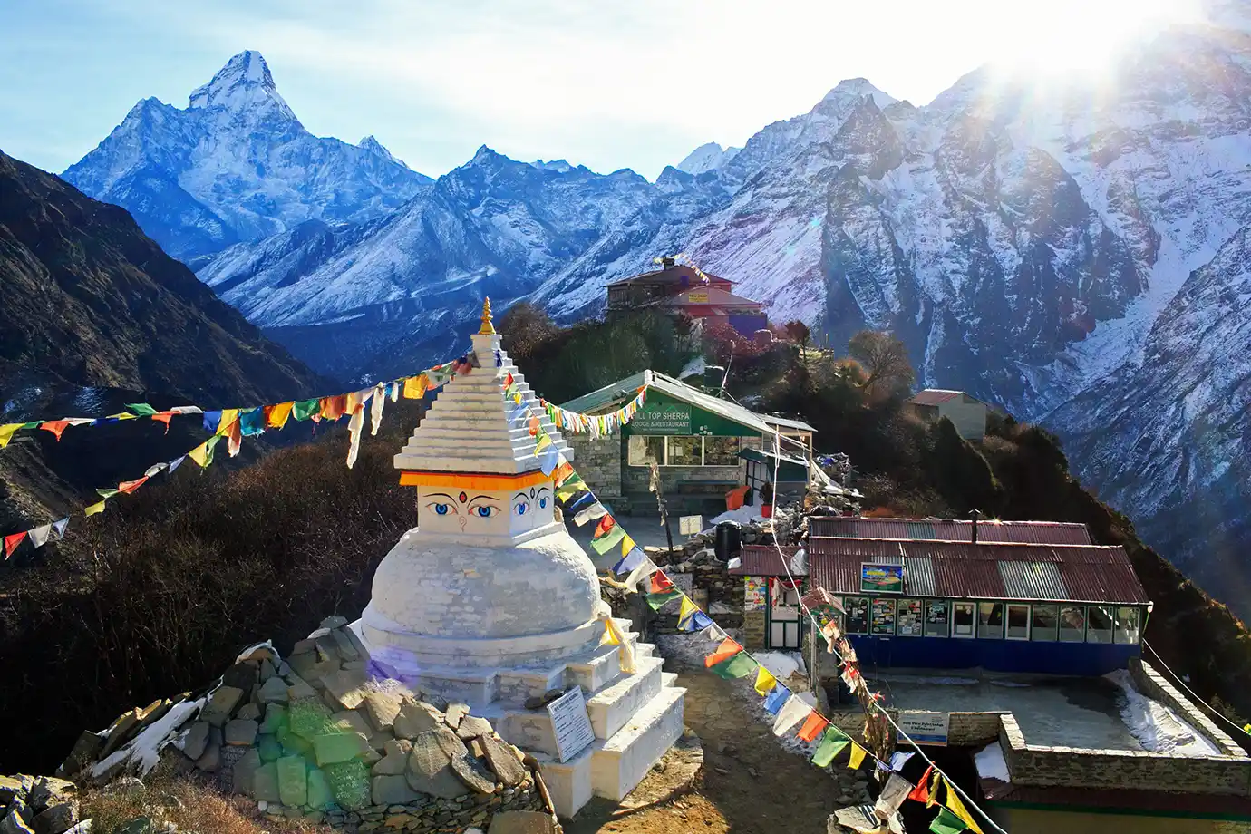 A wide high-angle landscape photograph showing a white Buddhist stupa draped with colorful prayer flags in the foreground, with stone lodges and the snow-capped peak of Mount Ama Dablam rising against a sunlit sky in Nepal.