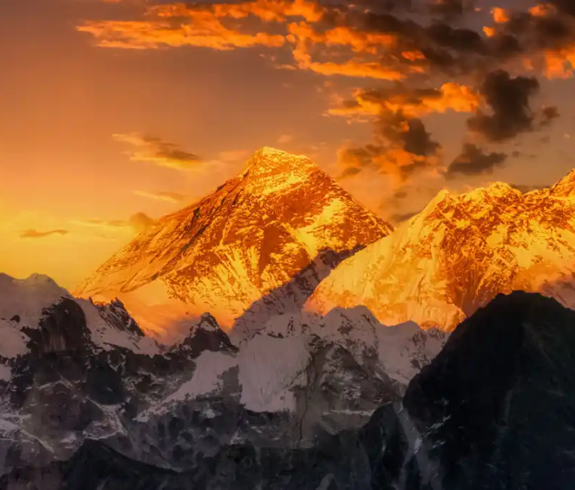 A dramatic wide landscape photograph of the snow-capped Himalayan peaks of Mount Everest and Lhotse illuminated by golden sunrise light under an orange and purple twilight sky in the Khumbu Region, Nepal.