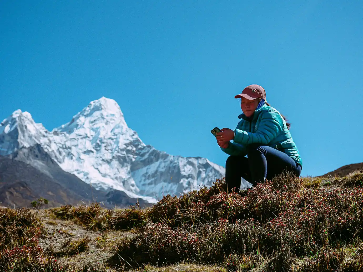 A low-angle outdoor portrait of a female trekker wearing a teal puffer jacket and cap, sitting on a grassy ridge while looking at her smartphone with the snow-covered peak of Mount Ama Dablam rising in the background in Nepal.