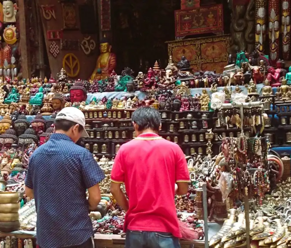 A wide eye-level photograph showing two men seen from behind browsing a dense display of traditional Nepalese handicrafts, brass singing bowls, carved wooden masks, and prayer wheels at a vibrant market stall in Thamel, Kathmandu.