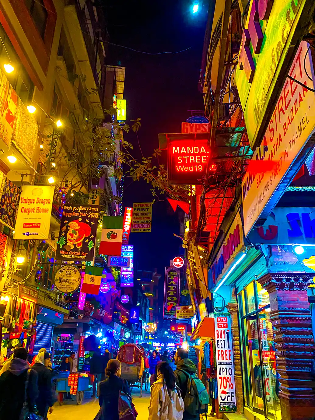 A low-angle vertical photograph of a narrow, brightly lit street at night in Thamel, Kathmandu, featuring glowing red, yellow, and blue neon signage, shop banners, overhead string lights, and pedestrians walking past commercial storefronts.   