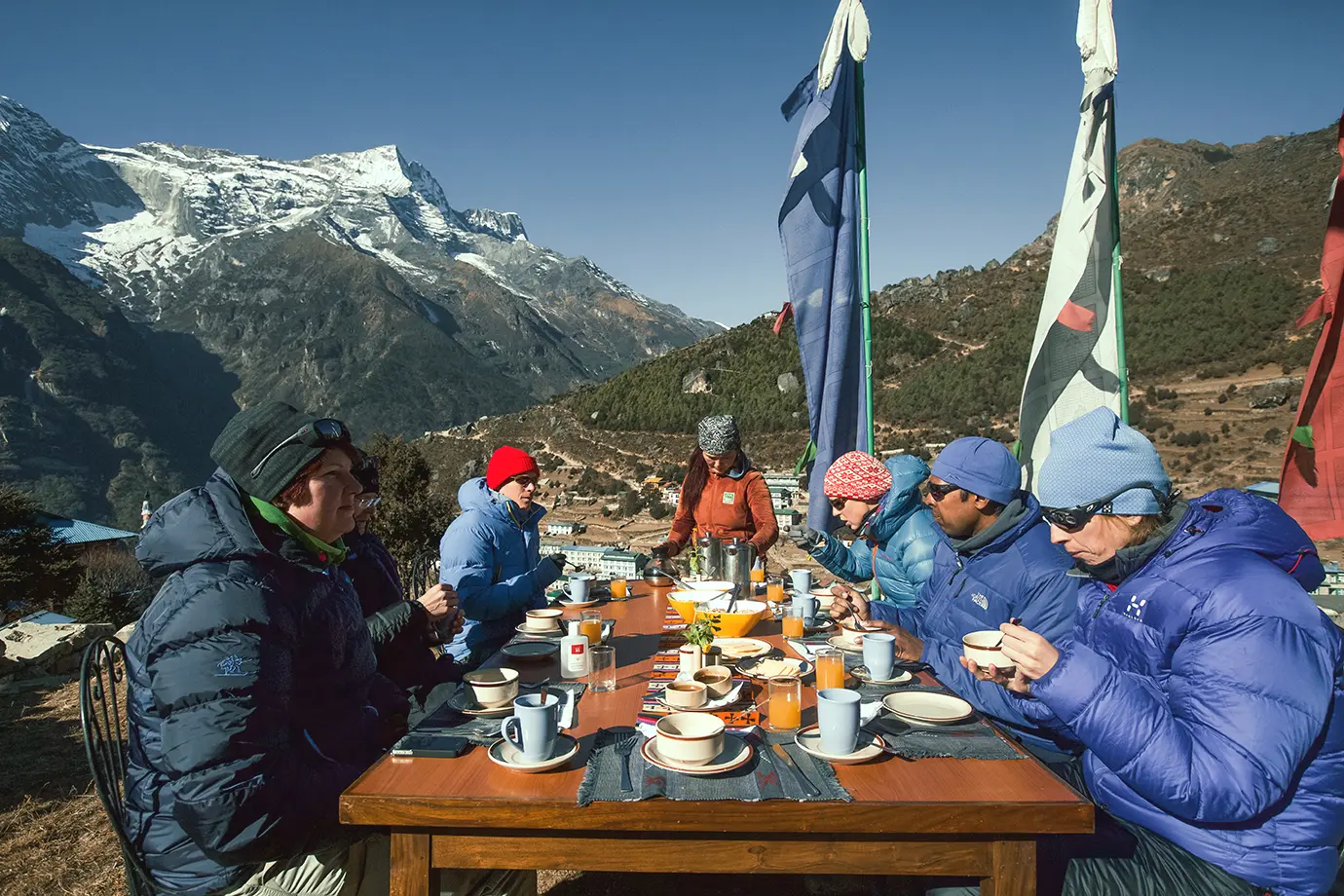 Trekkers enjoying breakfast outdoors at a long table with Himalayan mountains in the background and prayer flags nearby.