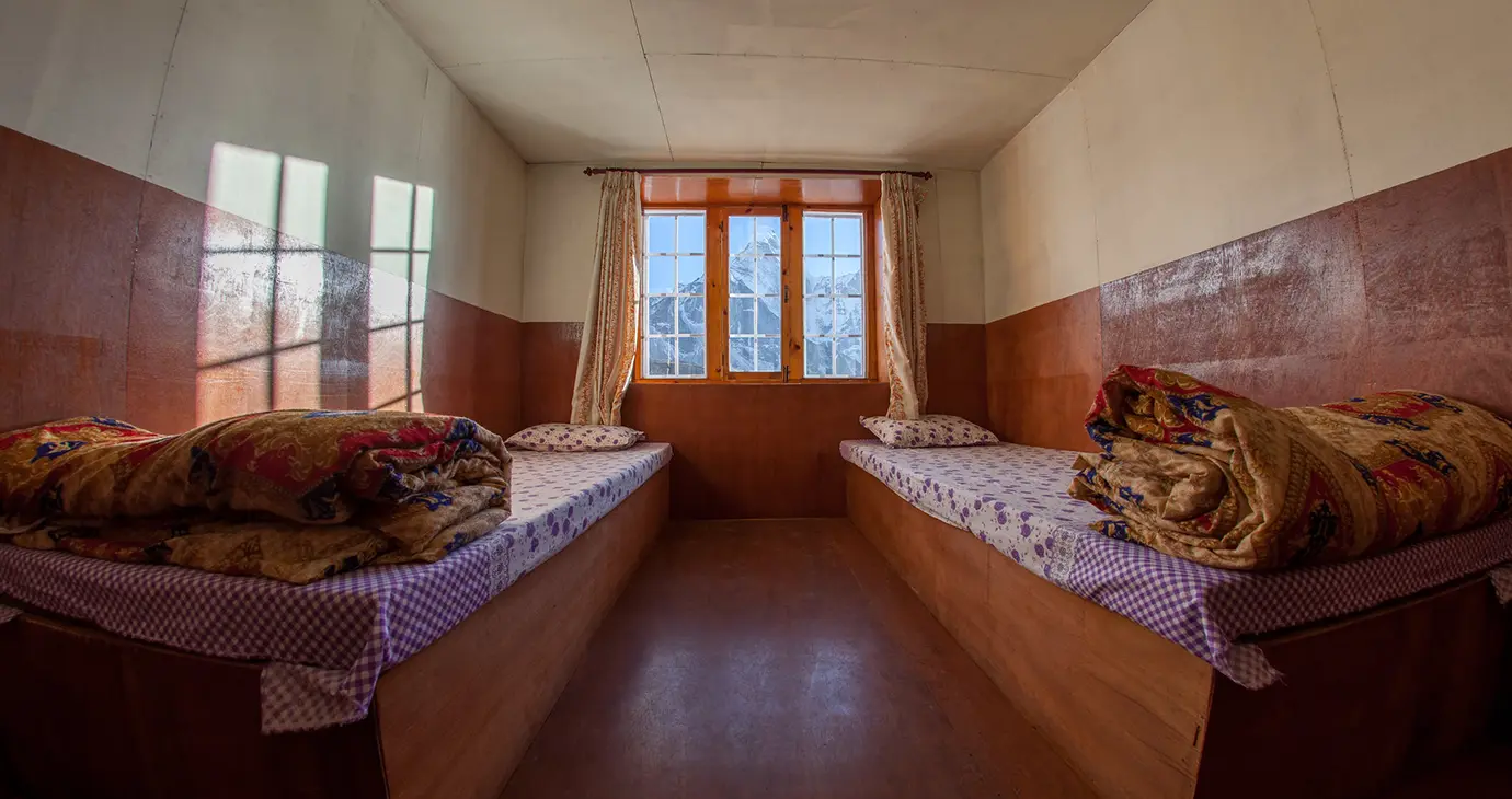 Simple twin room in a mountain teahouse with two single beds, folded blankets, and a window overlooking snow-covered Himalayan peaks.