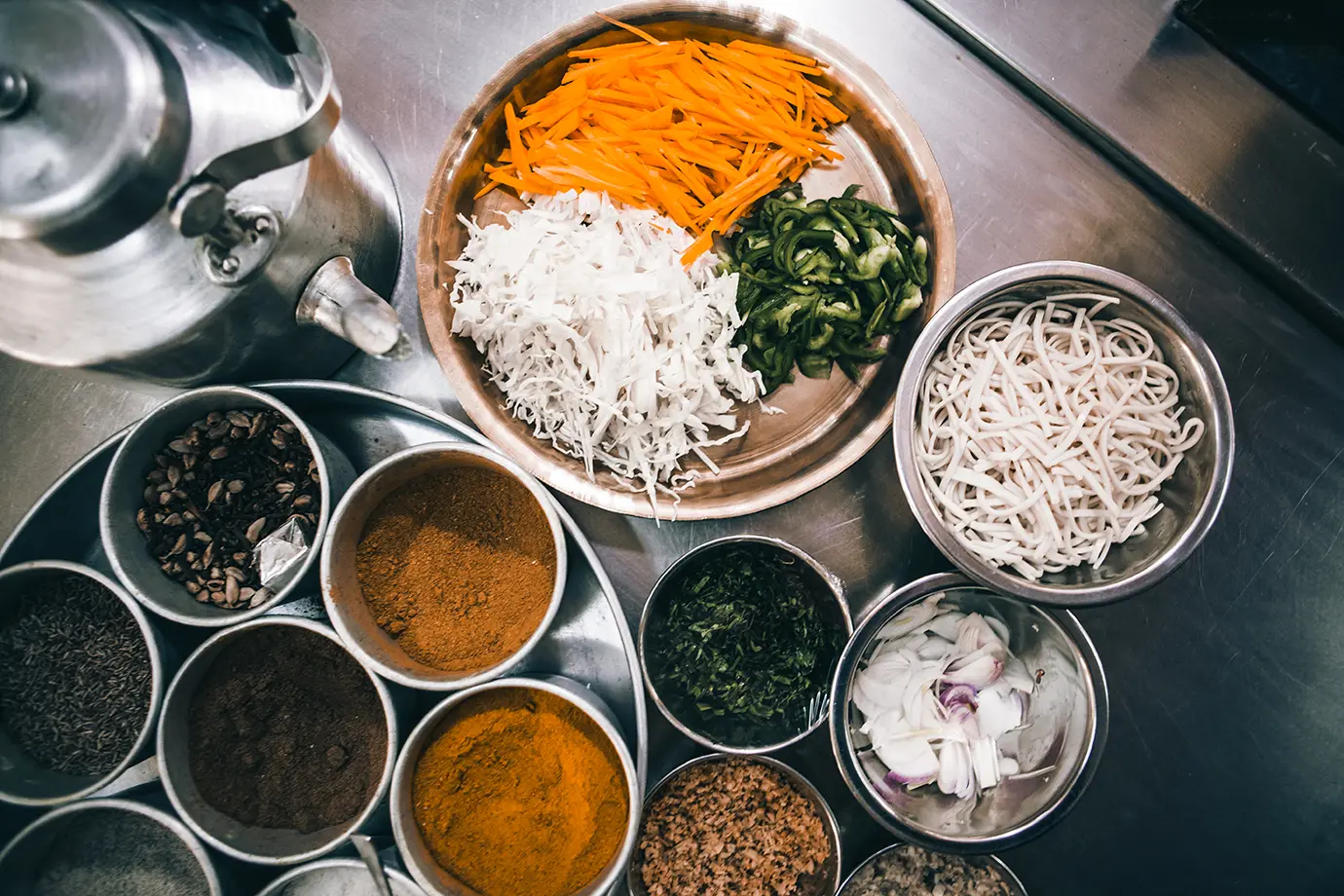 Freshly prepared vegetables, noodles, and assorted spices arranged in metal bowls in a Himalayan lodge kitchen.