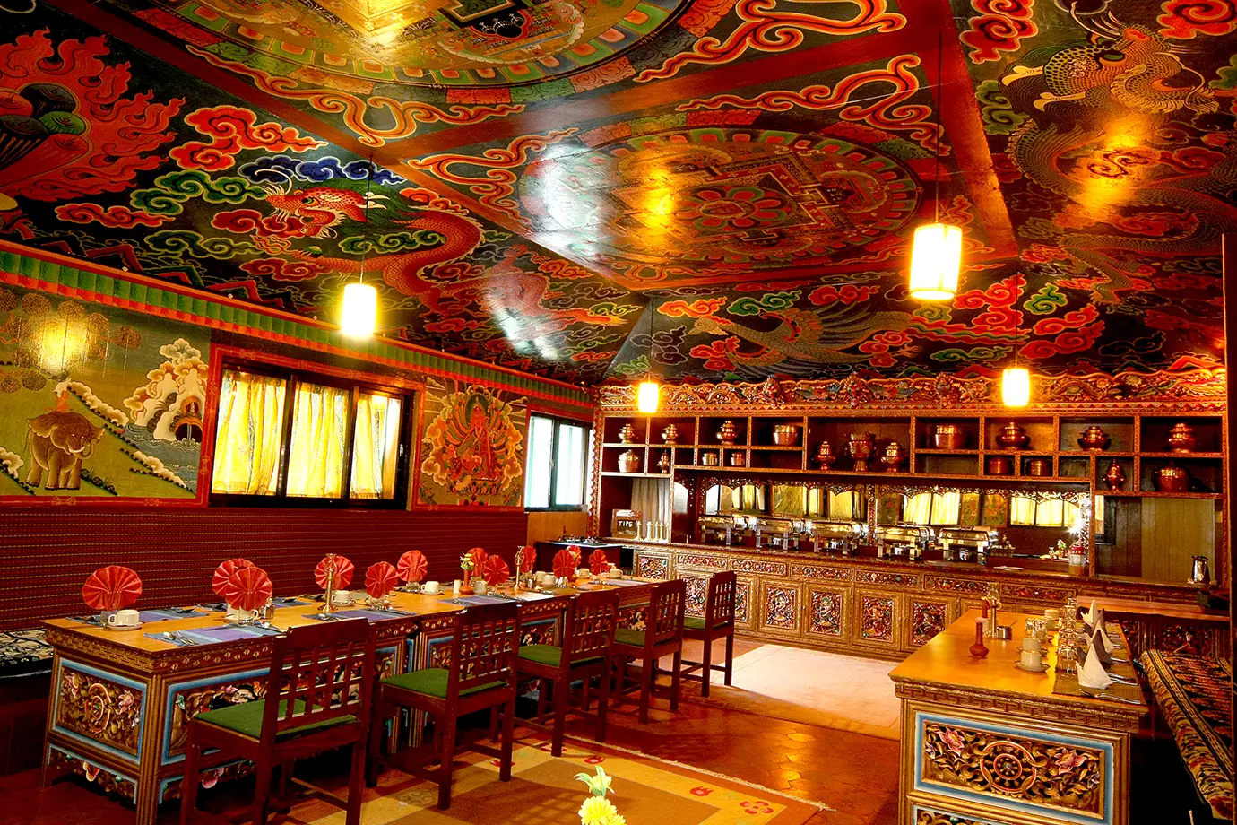 A colorful Tibetan-style dining hall with ornate wooden furniture, detailed wall murals, and a brightly painted ceiling.