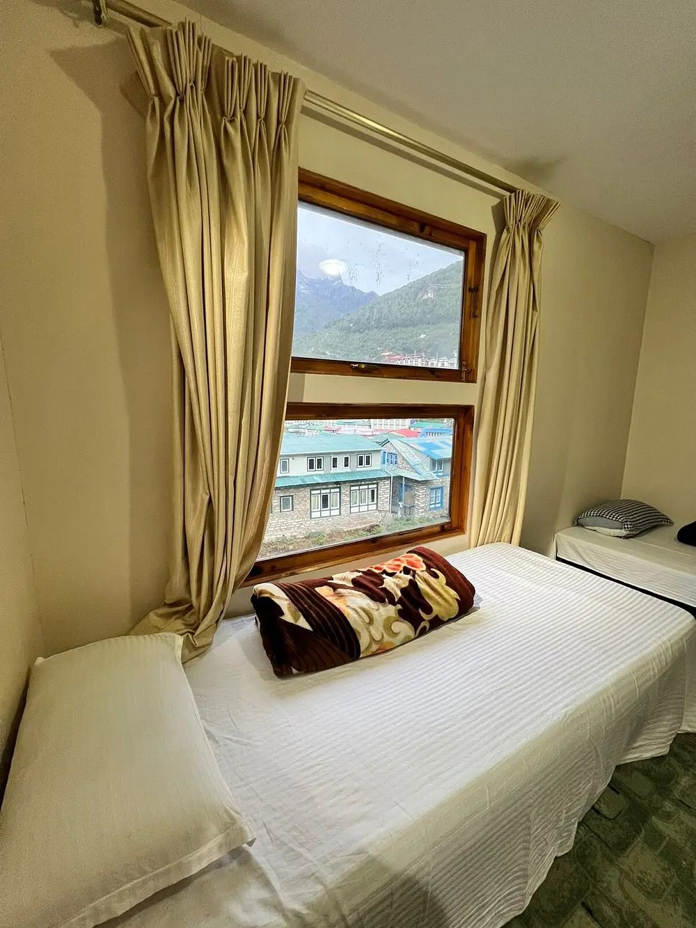 A simple twin room with neatly made beds, large windows, beige curtains, and a view of Namche Bazaar and surrounding hills.