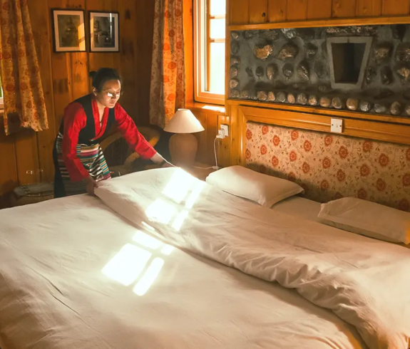 A lodge staff member neatly arranging bedding in a warm, wooden Himalayan guestroom with sunlight streaming through large windows.