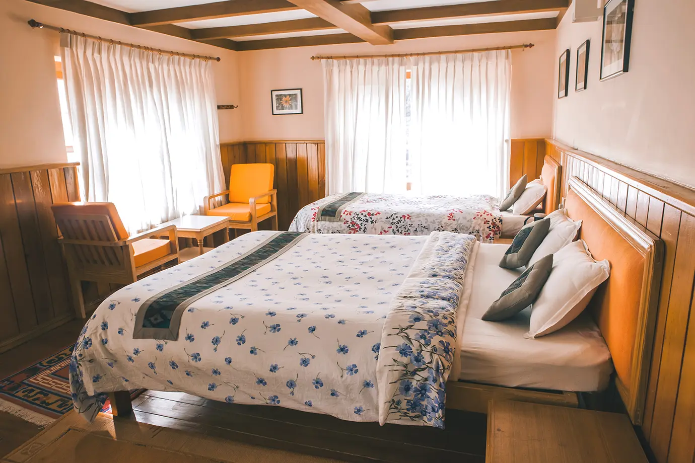 A bright and comfortable hotel room with two neatly made beds, wooden interiors, soft curtains, and a cozy seating area.
