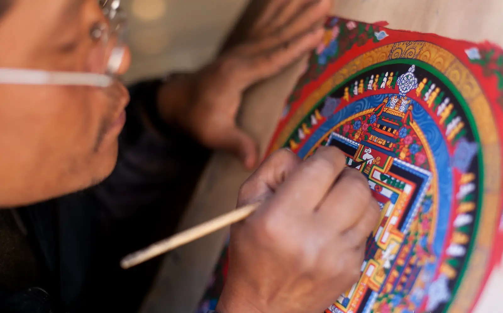 A close-up of an artist painting a traditional Tibetan Thangka, depicting a colorful mandala with intricate details.