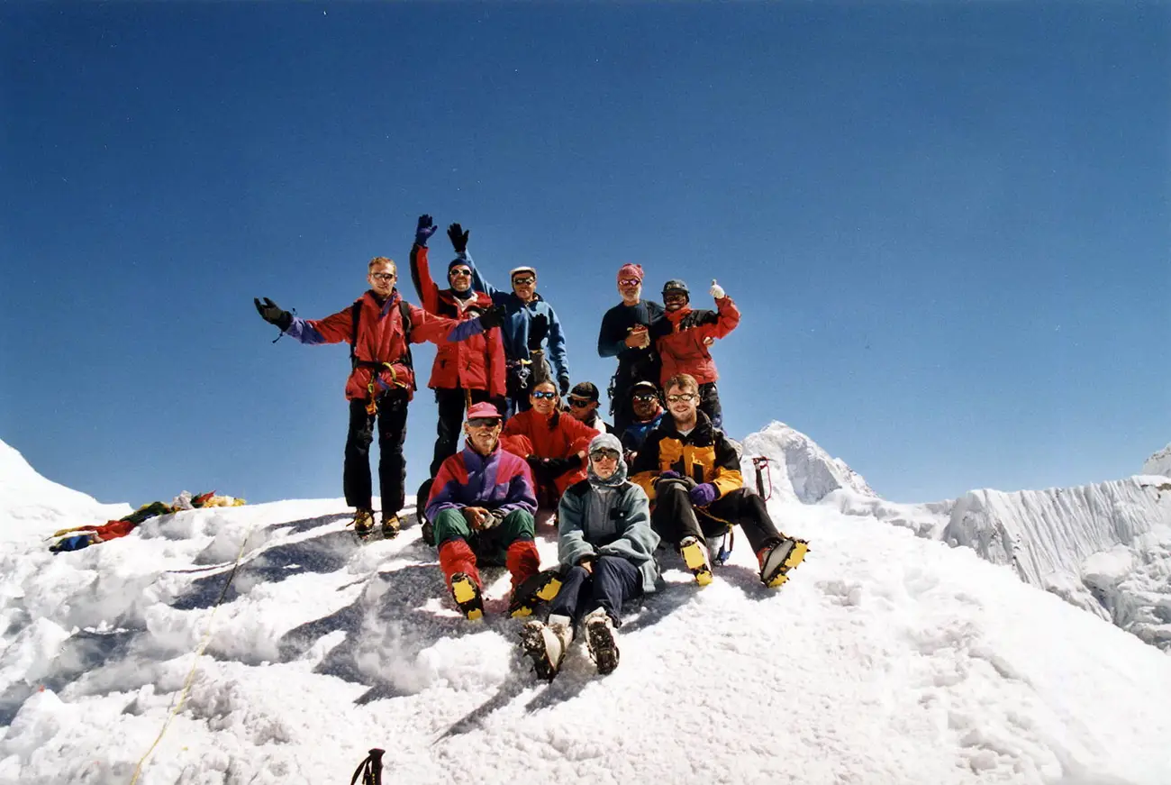 A group of climbers celebrating at the summit of Island Peak, wearing colorful mountaineering gear, surrounded by snow-covered peaks under a clear blue sky.
