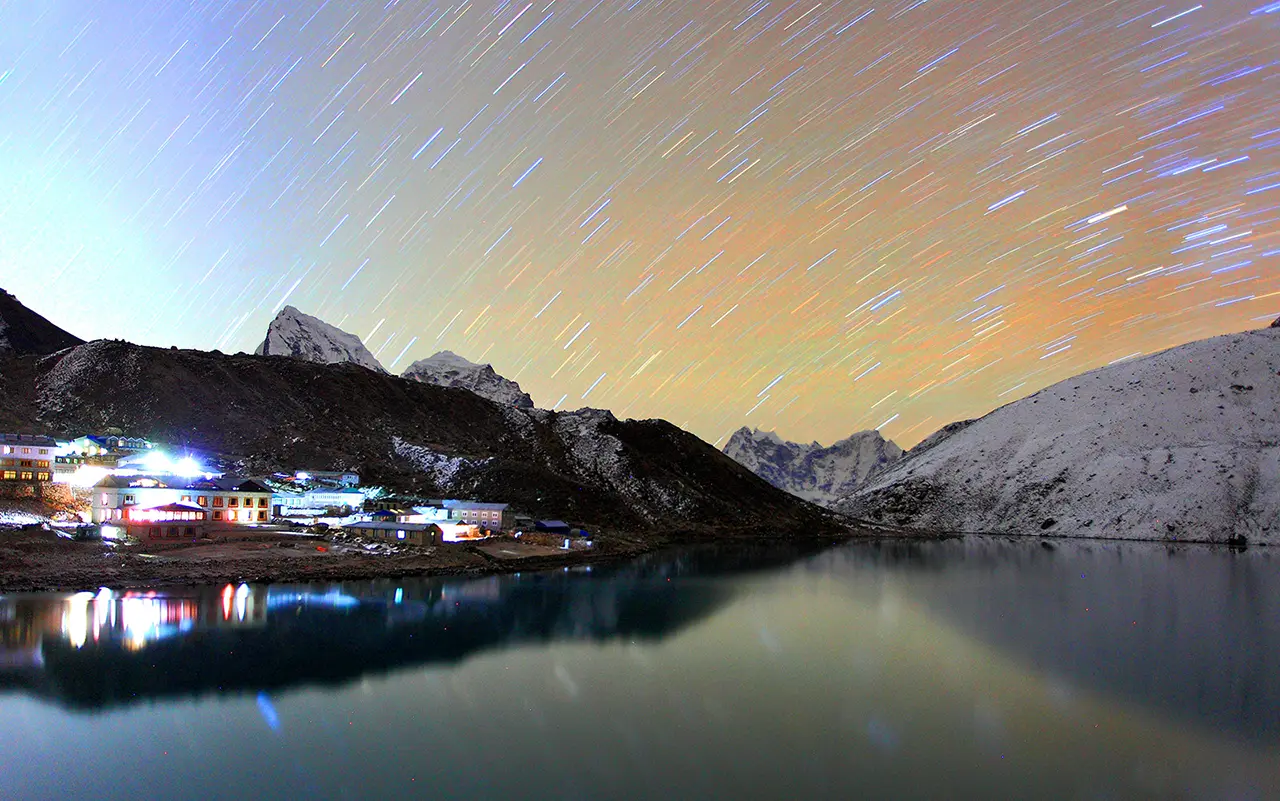 Những vệt sao trên bầu trời đêm hồ Gokyo, với hình bóng núi non và ánh đèn làng quê phản chiếu trên mặt nước tĩnh lặng.