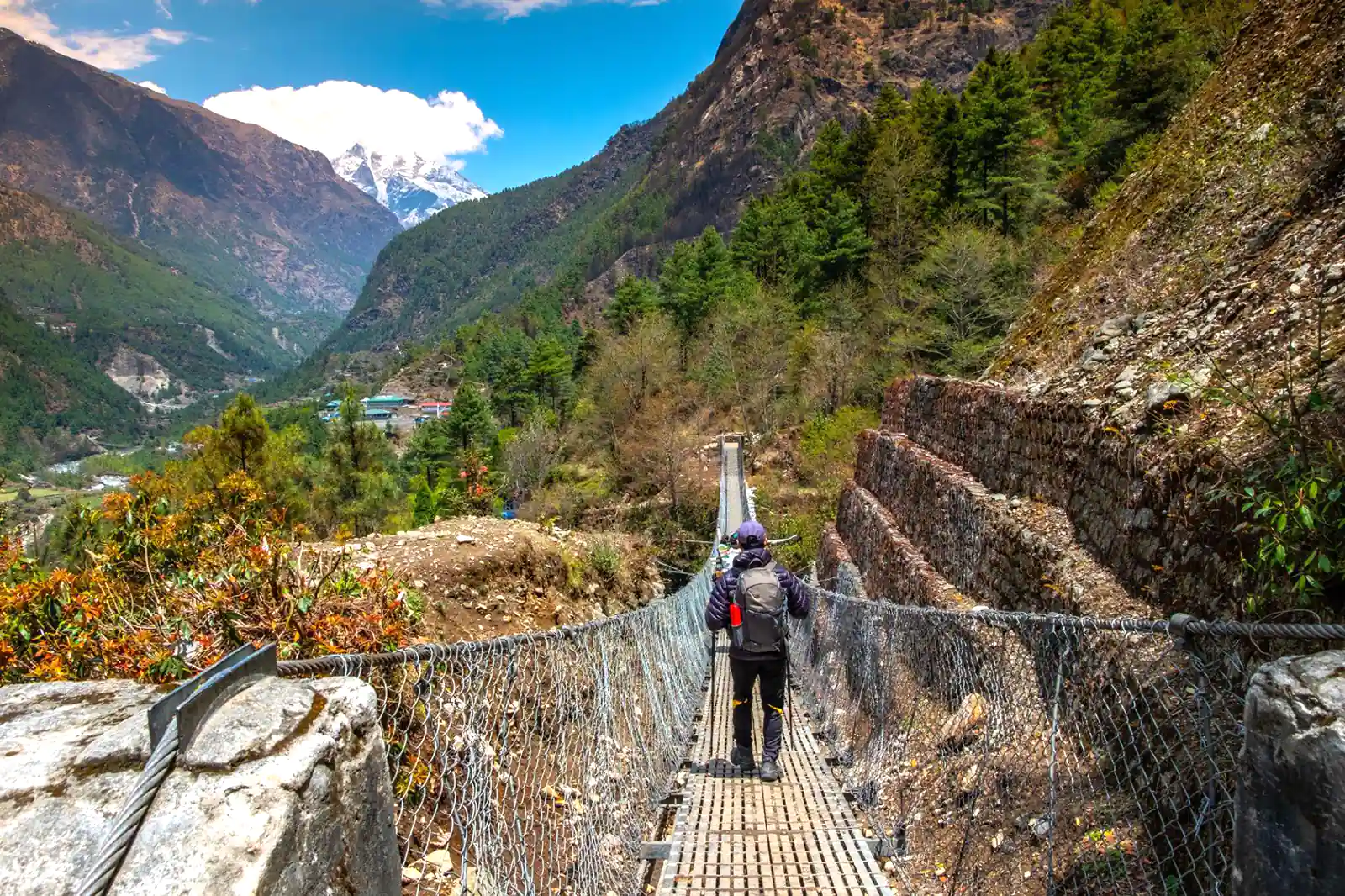 A backpacker crosses a suspension bridge on the trekking route from Lukla to Phakding, with snow-capped Himalayan mountains in the distance