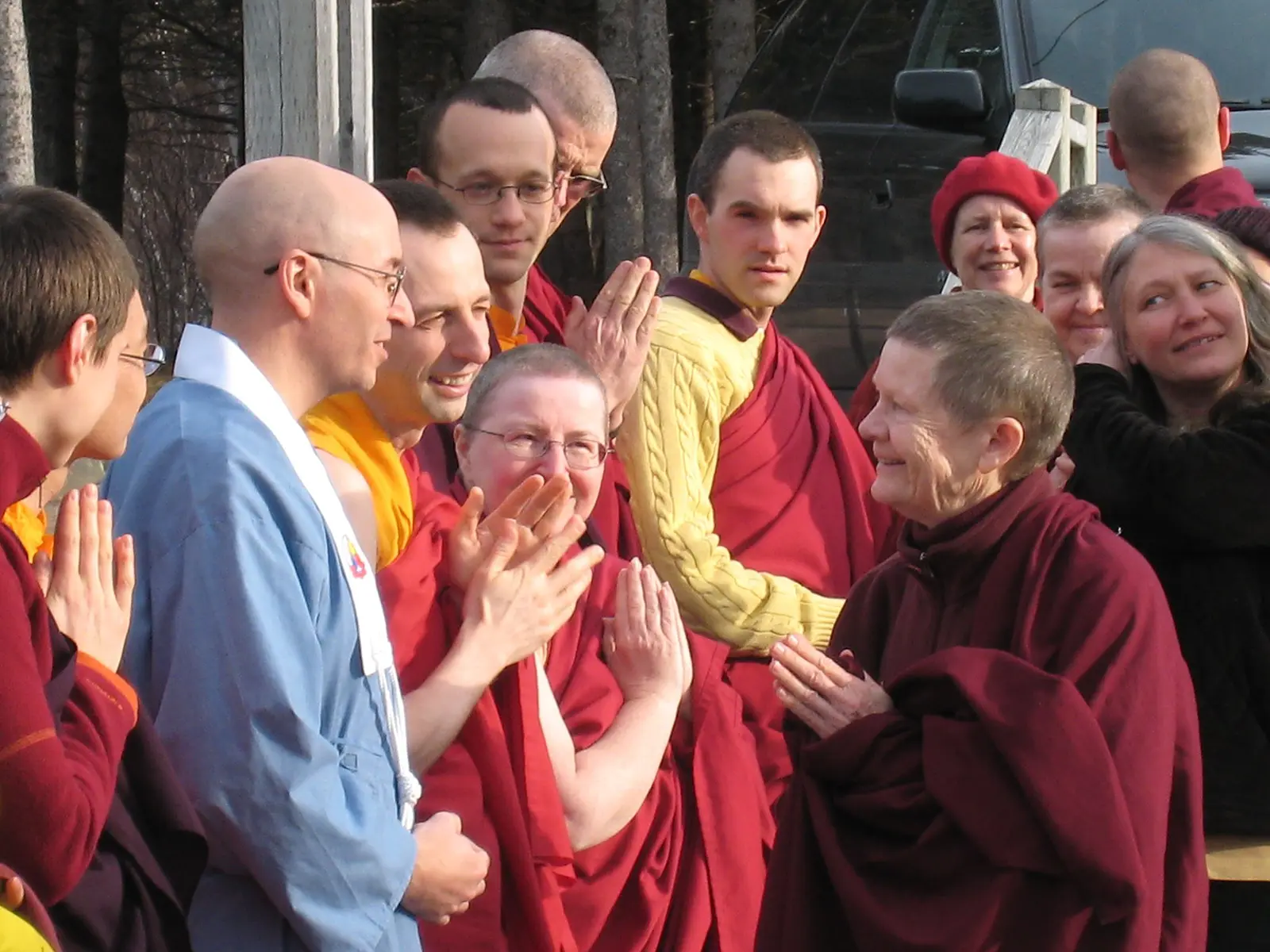 Pema Chödrön, dressed in maroon Buddhist robes, joyfully converses with a group of monks and nuns, all smiling and engaged.