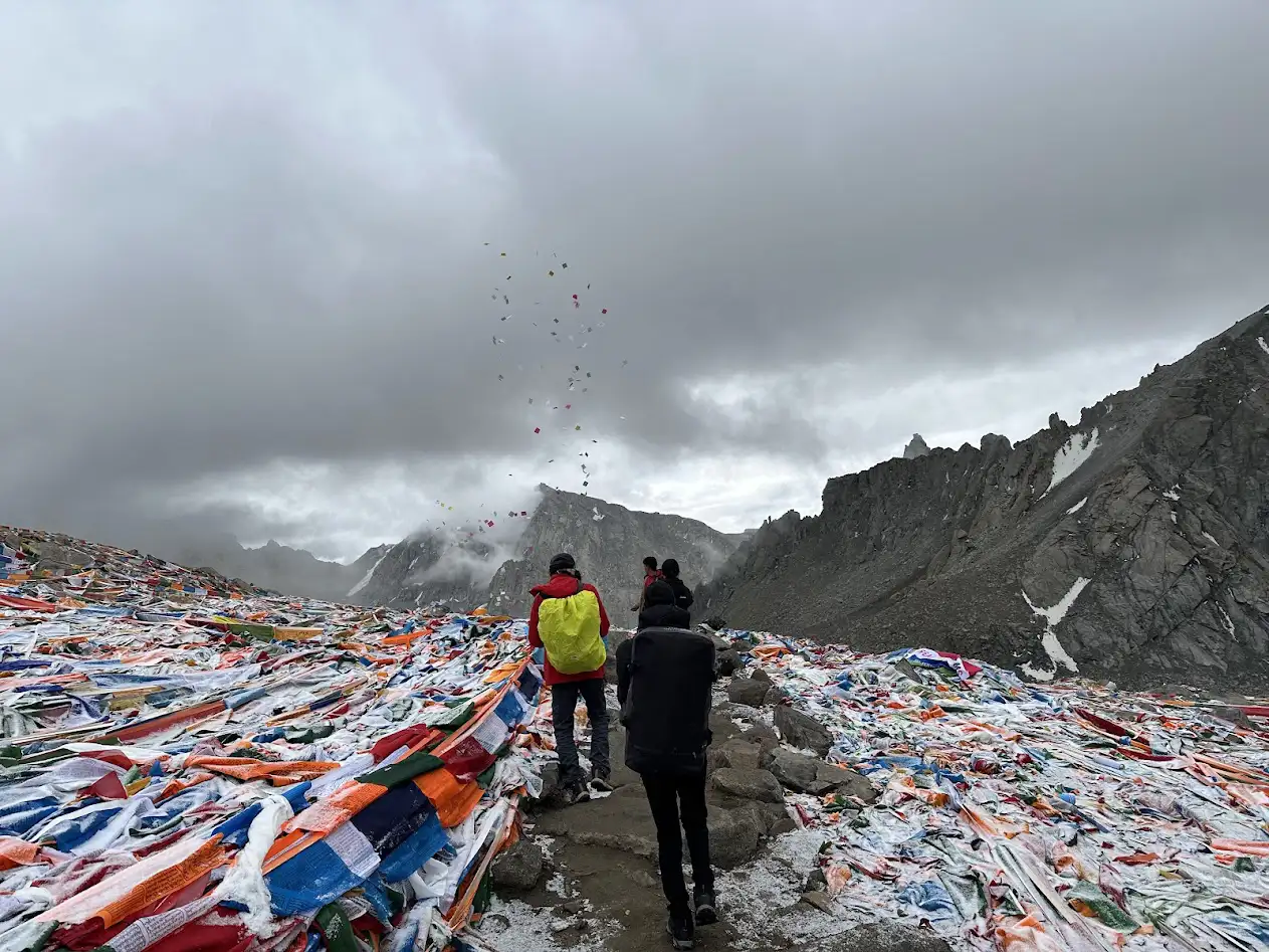 Travelers walking among dense prayer flags at Dolma La Pass, Tibet, with dark clouds overhead and rugged mountains in the background.