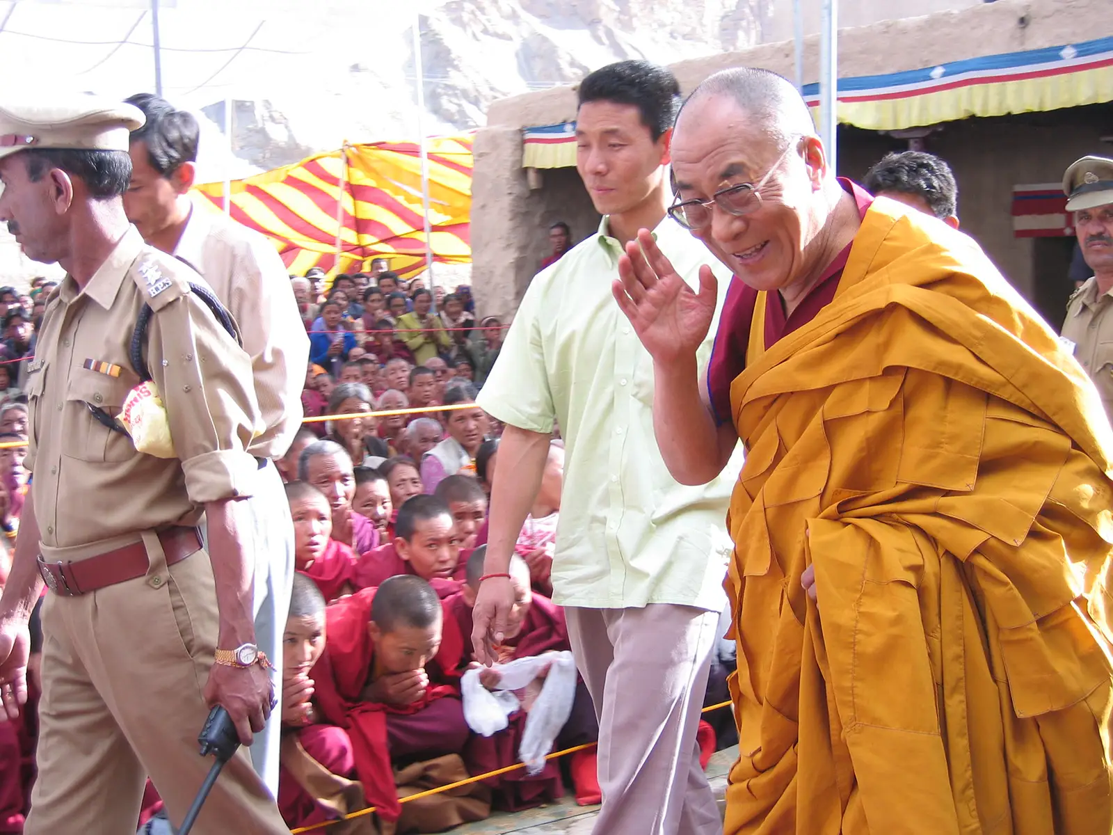 The Dalai Lama waving to a crowd during an event in Dharamsala, accompanied by security personnel.
