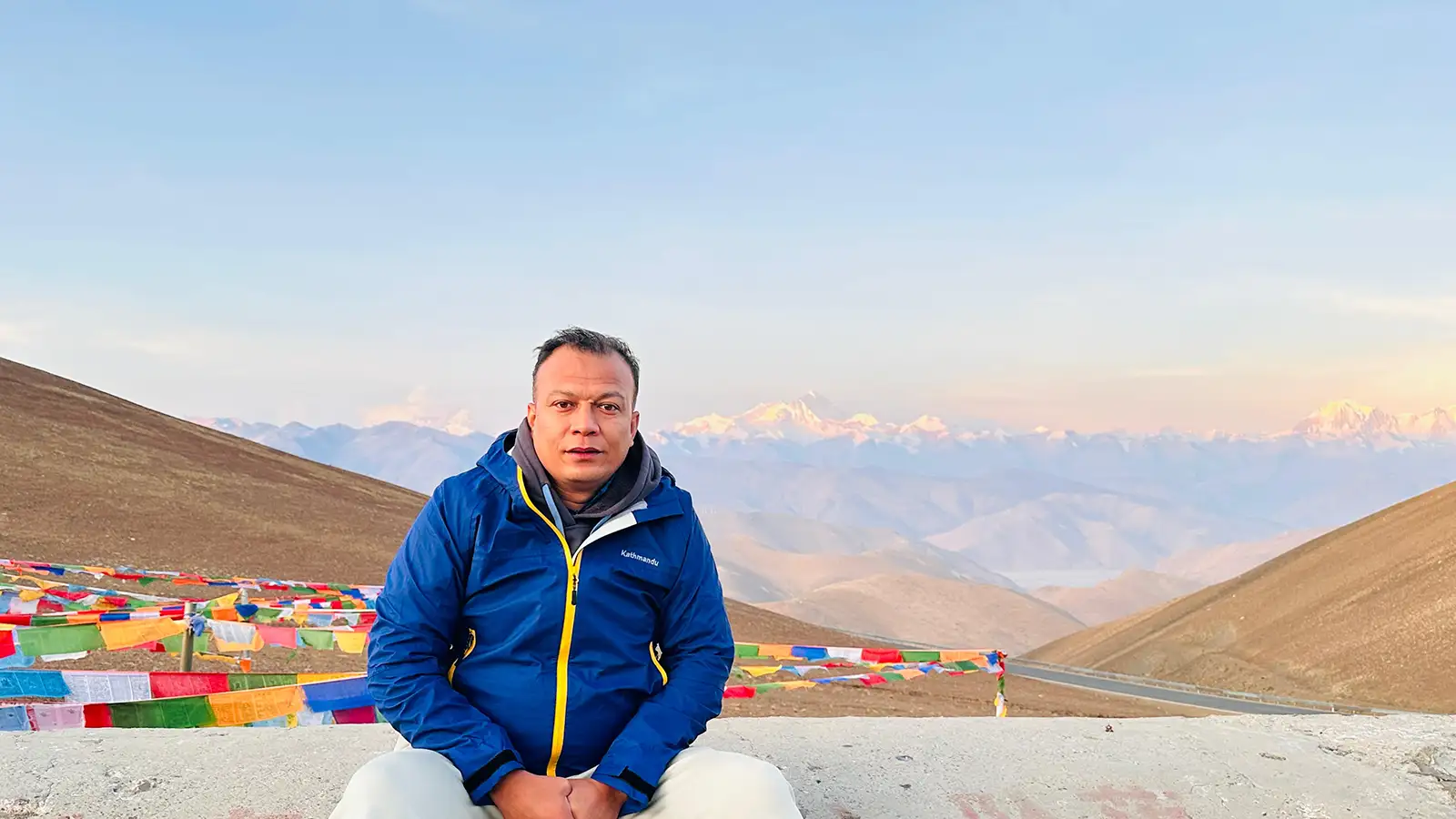A man in a blue jacket sitting in front of colorful prayer flags with a panoramic view of snow-capped mountains in the background.