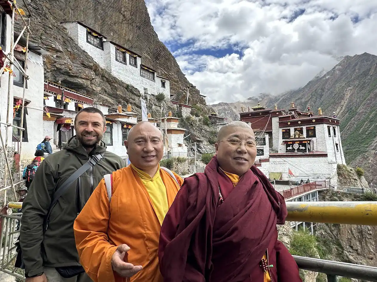 Zwei buddhistische Mönche und ein ausländischer Besucher lächeln gemeinsam im Höhlenkloster Milarepa, mit tibetischer Architektur im Hintergrund.
