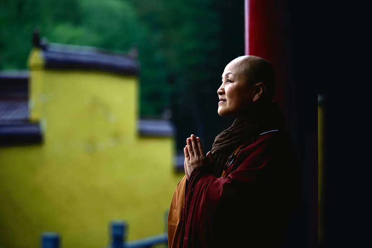 Pema Chödrön, dressed in traditional maroon Buddhist robes, stands in prayer with her hands together, looking serene with a yellow wall and greenery in the background.