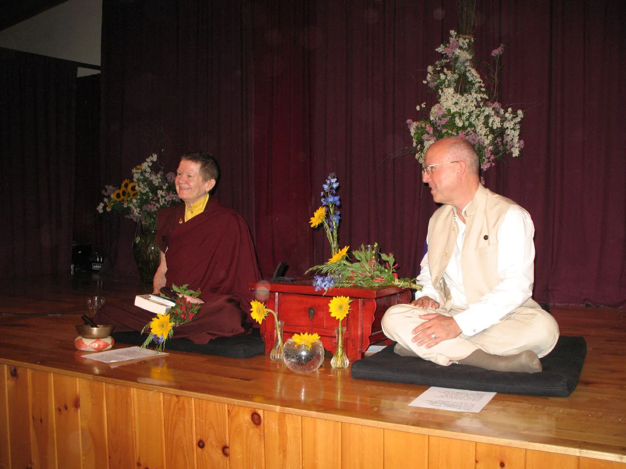 Pema Chödrön, dressed in maroon Buddhist robes, and Richard Reoch, in a light-colored suit, sit on a stage with flowers and a small table between them, both smiling.
