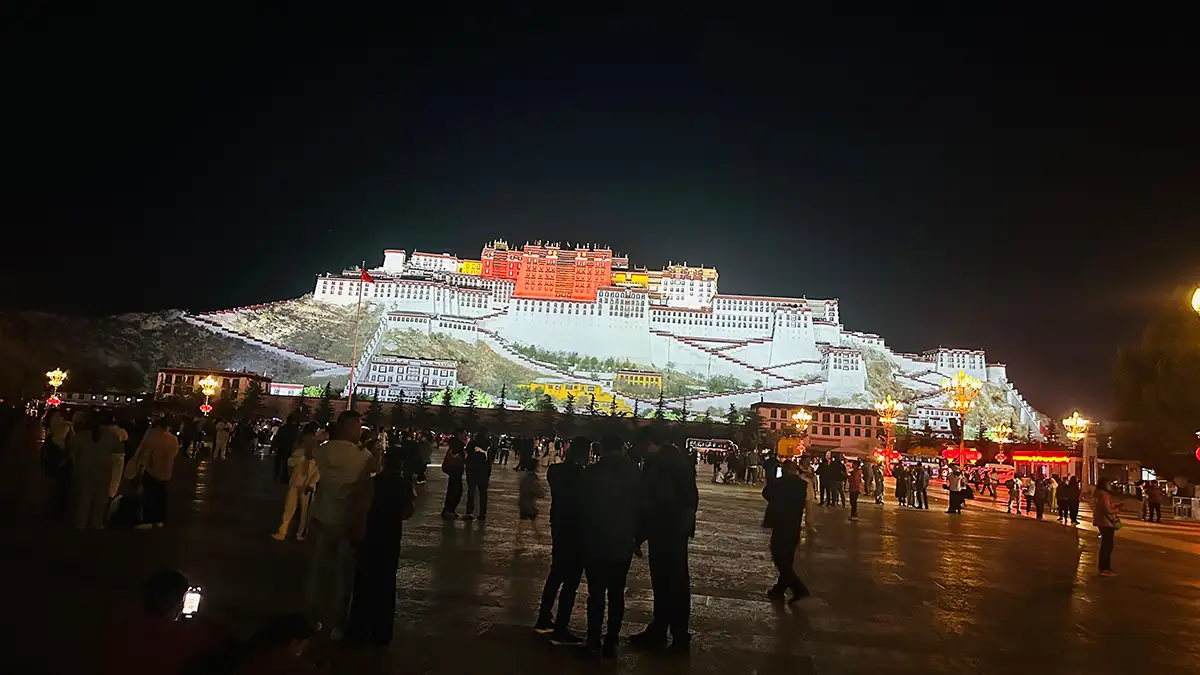 Night view of the illuminated Potala Palace in Lhasa, Tibet, with a crowd of people gathered in the foreground.
