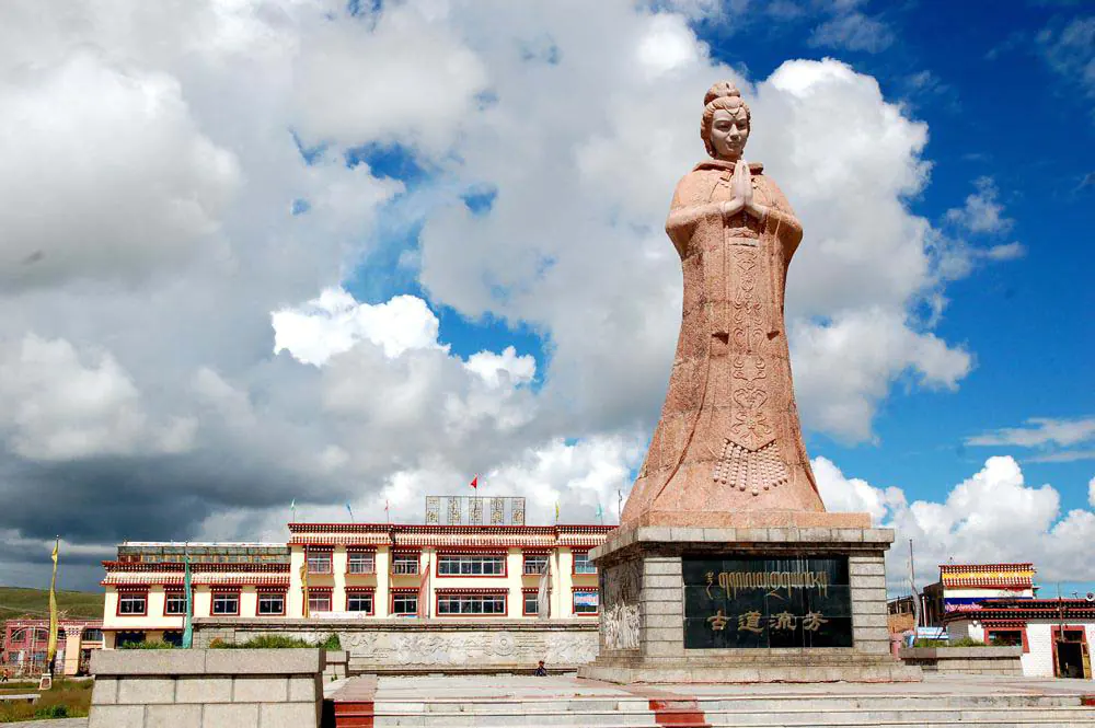Large stone statue of Princess Wencheng with hands clasped in prayer, set against a backdrop of a partly cloudy sky and traditional Tibetan buildings in Daotanghe, Qinghai.