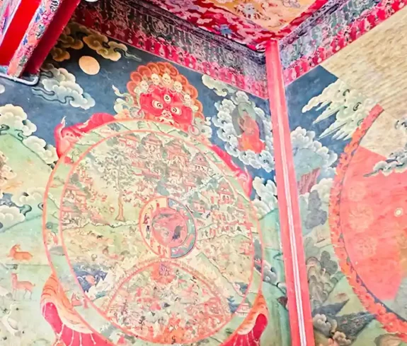 A vibrant mural of the Bhavacakra, or Wheel of Life, adorns a wall in a Tibetan Buddhist monastery. A monk dressed in red robes sits in the foreground, contemplating the intricate artwork.
