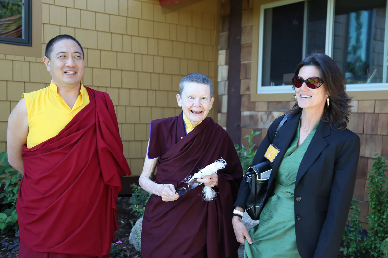 Shechen Rabjam Rinpoche, dressed in maroon and yellow robes, Ani Pema Chödrön in maroon robes, and Elizabeth Mattis Namgyel in a green dress and black blazer, smiling and waiting outside a building.