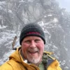 A man in a yellow jacket and black beanie smiling with snow-covered Taktsang Monastery in the background.