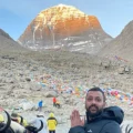 A man standing with hands in prayer pose in front of Mount Kailash at Tibet altitude, adorned with prayer flags and fellow travelers in the background.