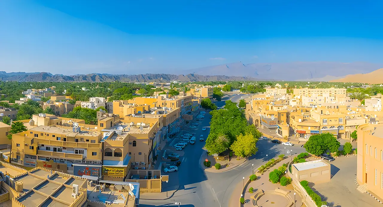 Aerial view of Nizwa town taken from the top of the local fortress in Oman, with traditional buildings, greenery, and distant mountains.