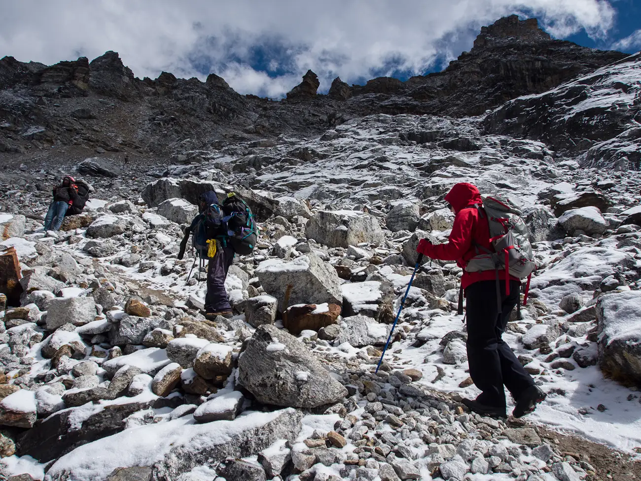 Three hikers ascending the rocky, snow-dusted terrain of Renjo La Pass with cloud-shrouded peaks in the background.