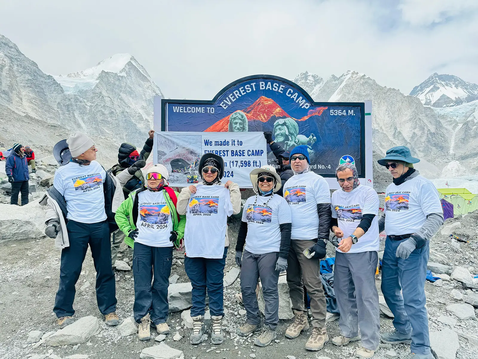 Group of seven hikers in matching t-shirts posing proudly at Everest Base Camp with a banner and mountains in the background.