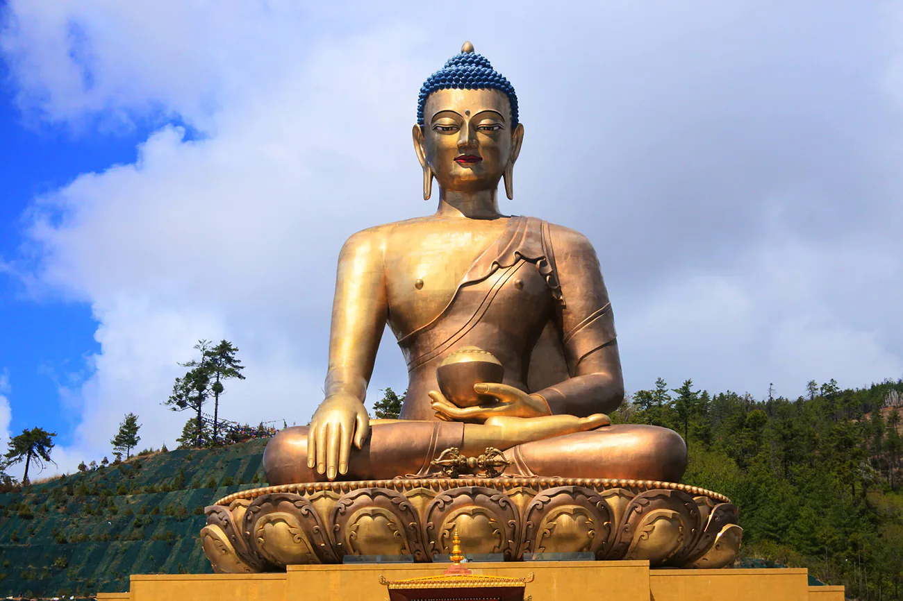 A colossal, golden Buddha Dordenma statue seated in the lotus position under a clear blue sky, surrounded by verdant hills.