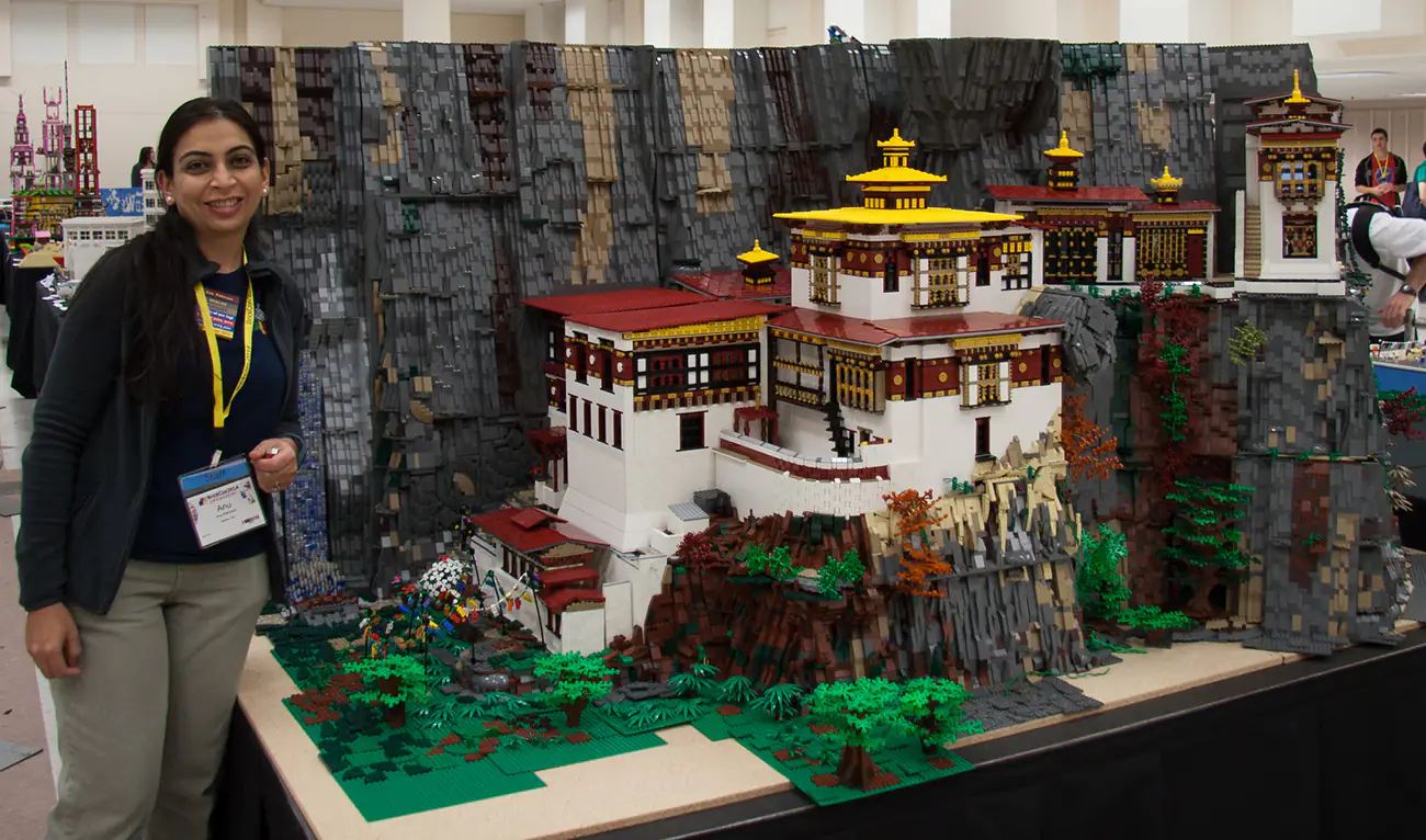 A woman proudly stands beside a detailed Lego model of the Taktsang Monastery, also known as Tiger's Nest, displayed at a public exhibition.