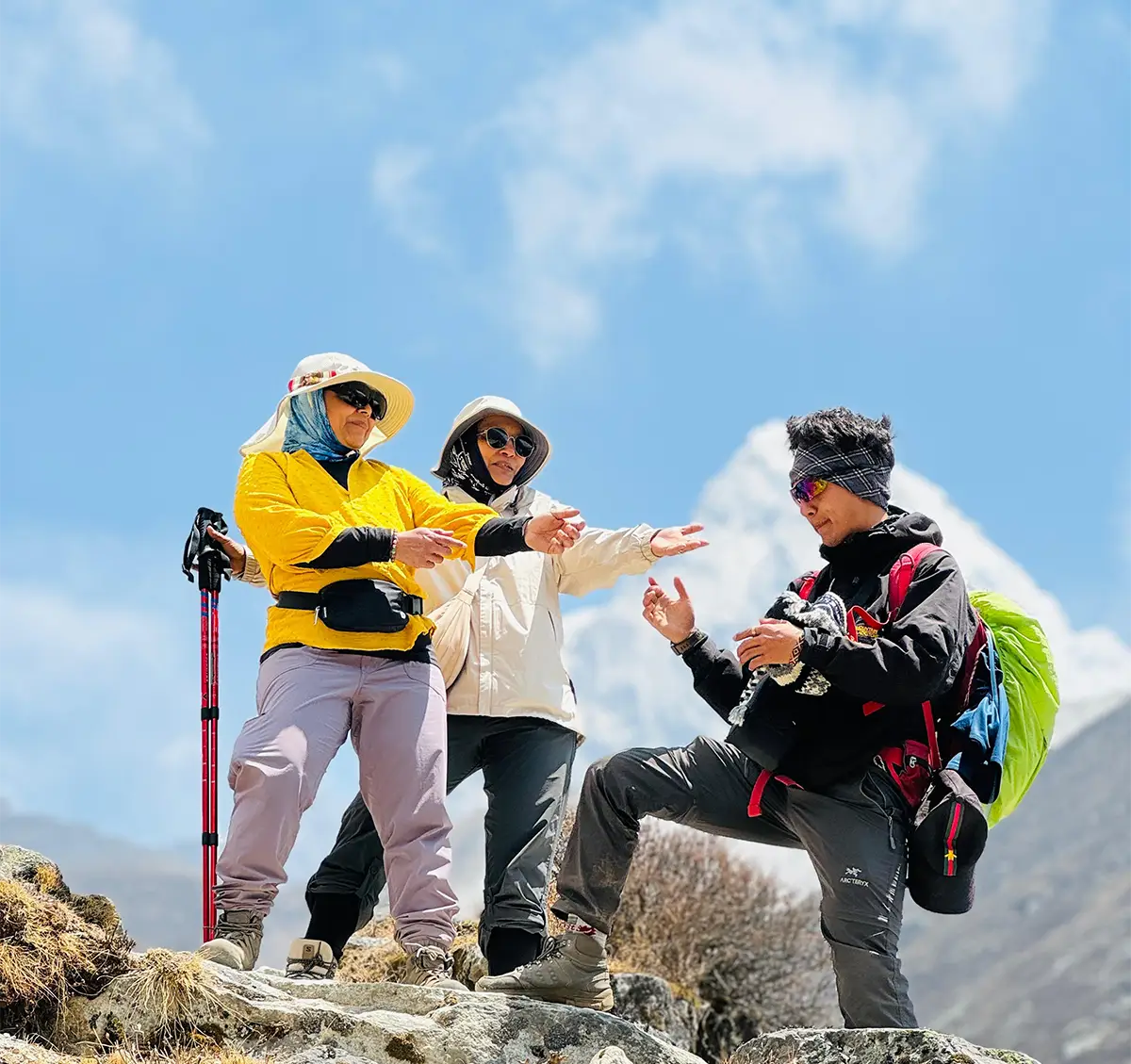 Three hikers in colorful outdoor gear interacting on a rocky path with a clear blue sky and mountain in the background during a trek.