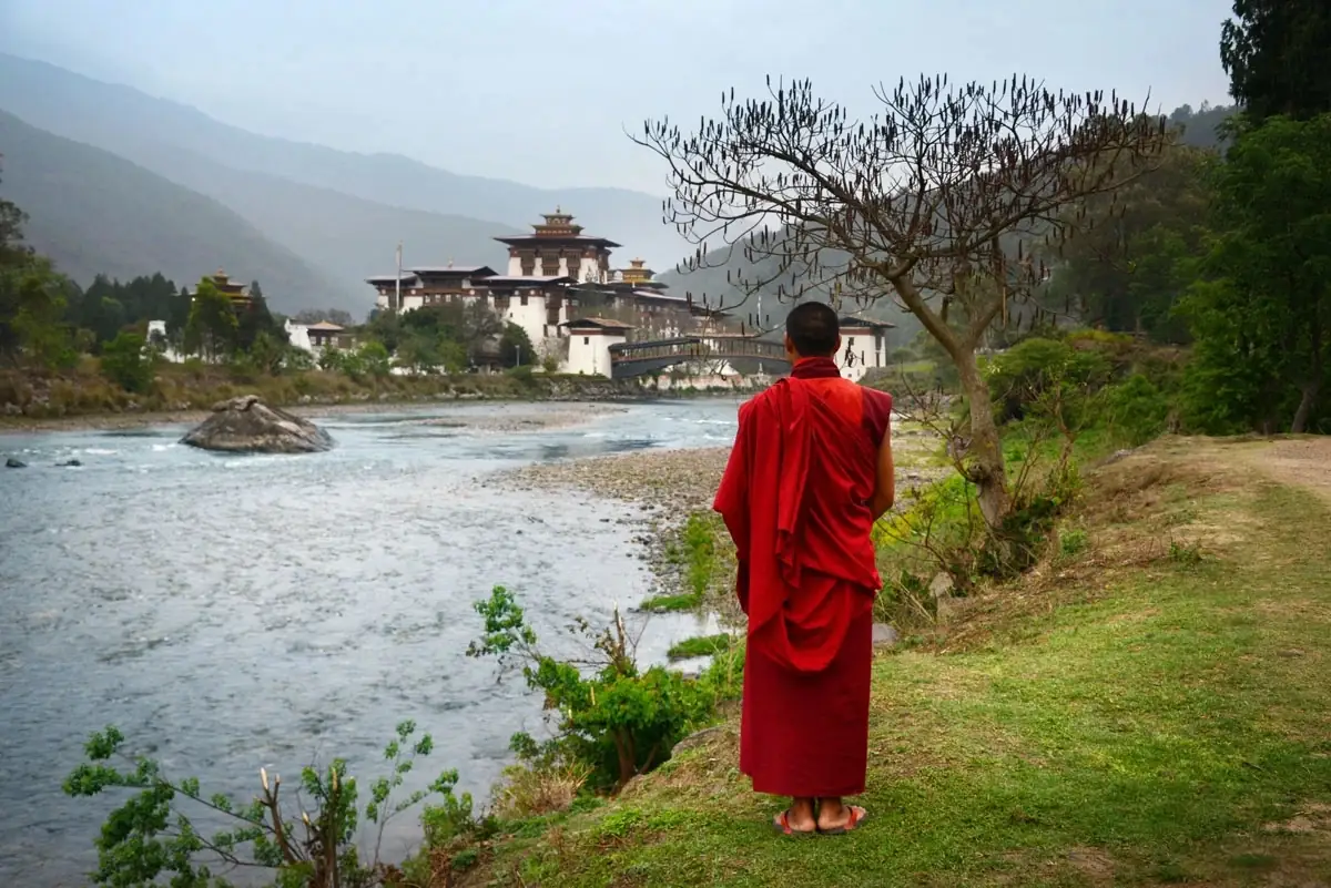 A Buddhist monk in a red robe standing by the river, gazing at Punakha Dzong in Bhutan, surrounded by lush greenery and flowing waters.