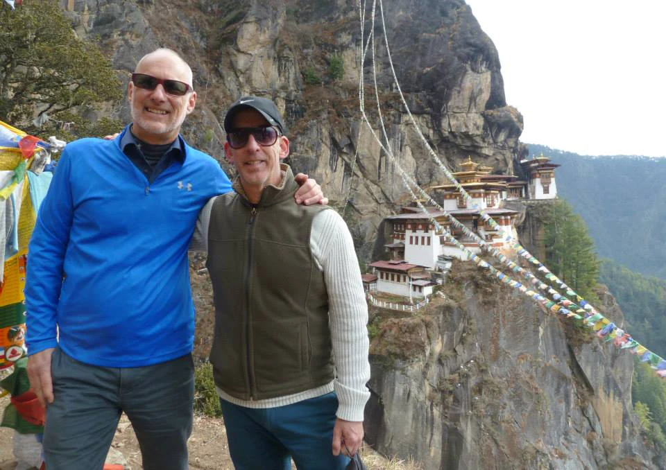Two men smiling, standing in front of the Tiger's Nest Monastery in Bhutan, with prayer flags and the iconic cliffside monastery in the background.