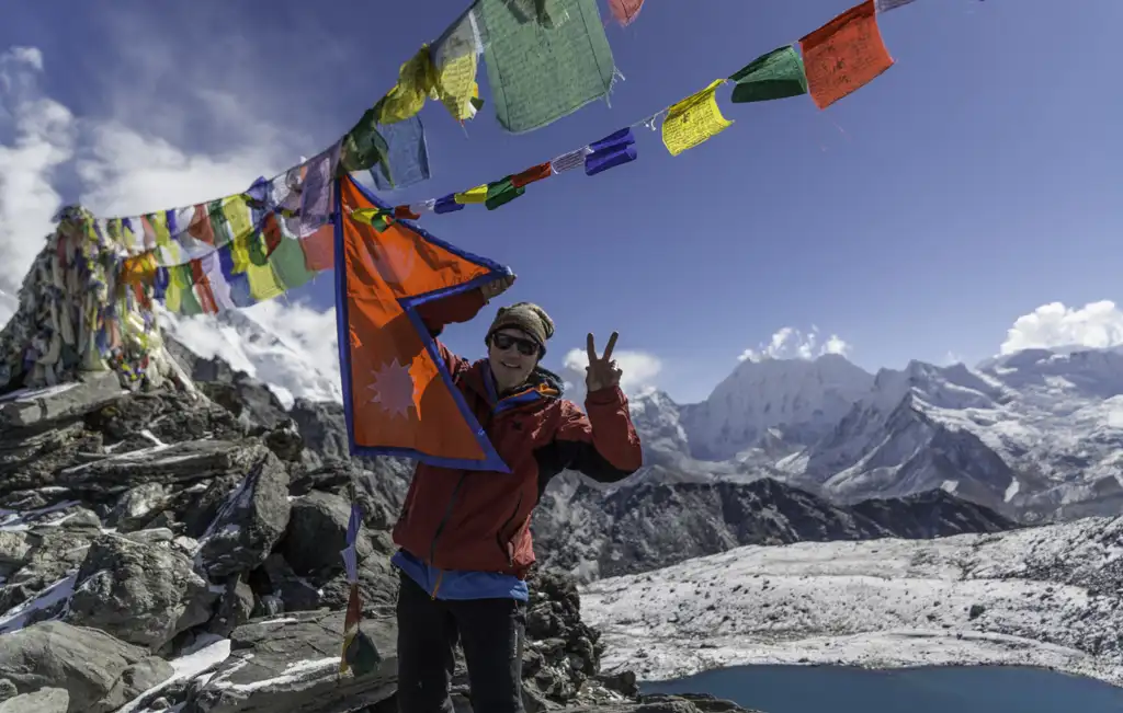 A hiker displaying a peace sign and holding a Nepalese flag at Kongma La Pass, surrounded by colorful prayer flags and snow-capped mountains.