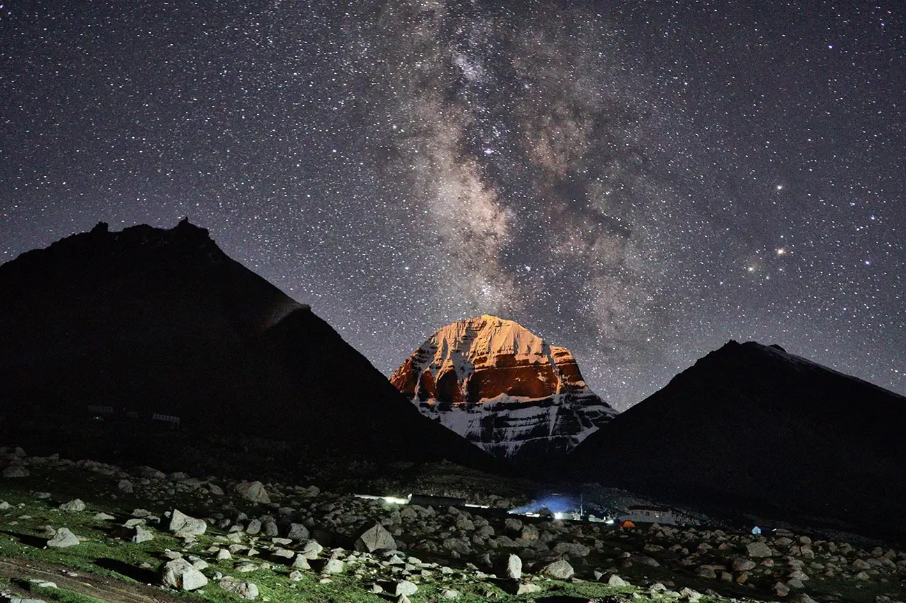 Mount Kailash illuminated under a starry night sky with the Milky Way visible above, surrounded by dark silhouettes of adjacent peaks and a rocky foreground.