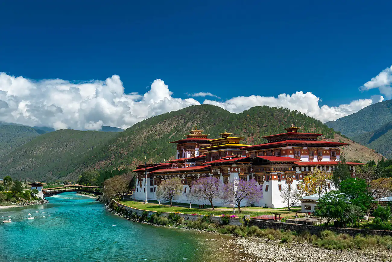 The Punakha Dzong, a majestic traditional Bhutanese fortress, sits by the side of a clear turquoise river, with a backdrop of lush green mountains and a bright blue sky.