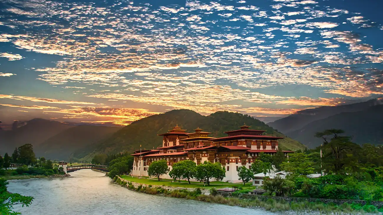 Stunning sunset view of Punakha Dzong in Bhutan, with the dzong illuminated by the golden light of the setting sun, the Mo Chhu river flowing in the foreground, and a sky dotted with scattered clouds.