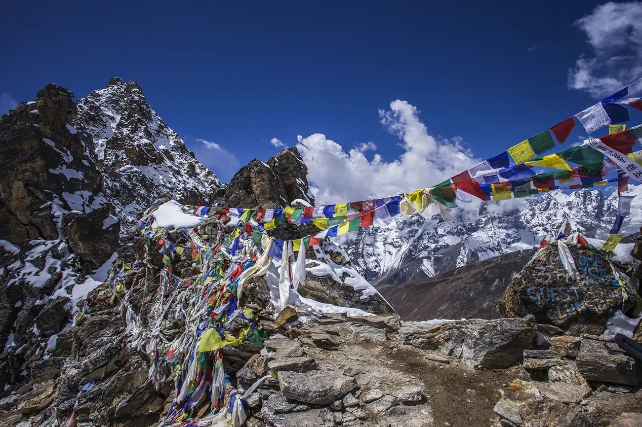 Colorful prayer flags strung across a rocky pass with snow-capped peaks in the background under a vivid blue sky.