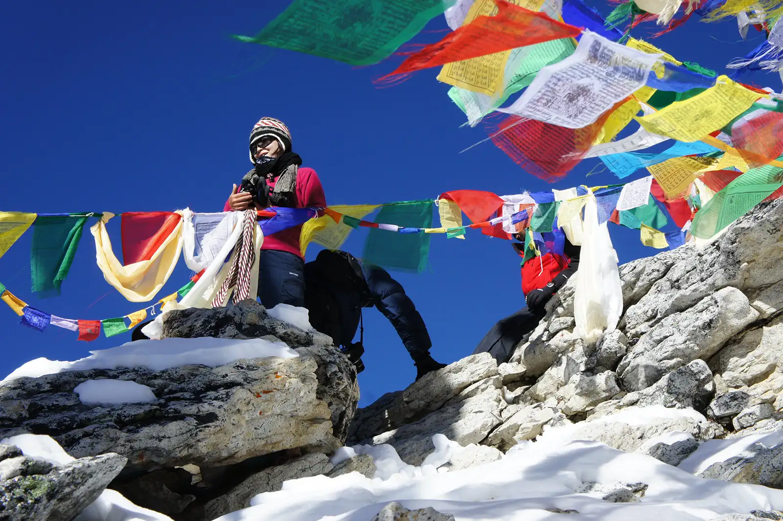 A solo trekker adjusting a camera, standing on a rocky outcrop adorned with colorful prayer flags, with a clear blue sky above in the Himalayas.