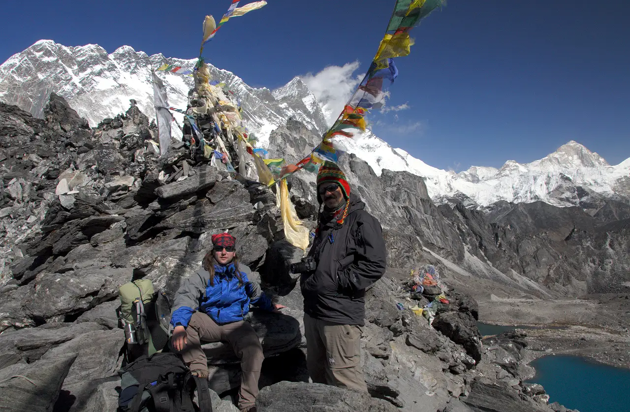 Two hikers seated among colorful prayer flags at the summit of Kongma La Pass, with a panoramic view of snow-capped mountains and a turquoise lake in the background.